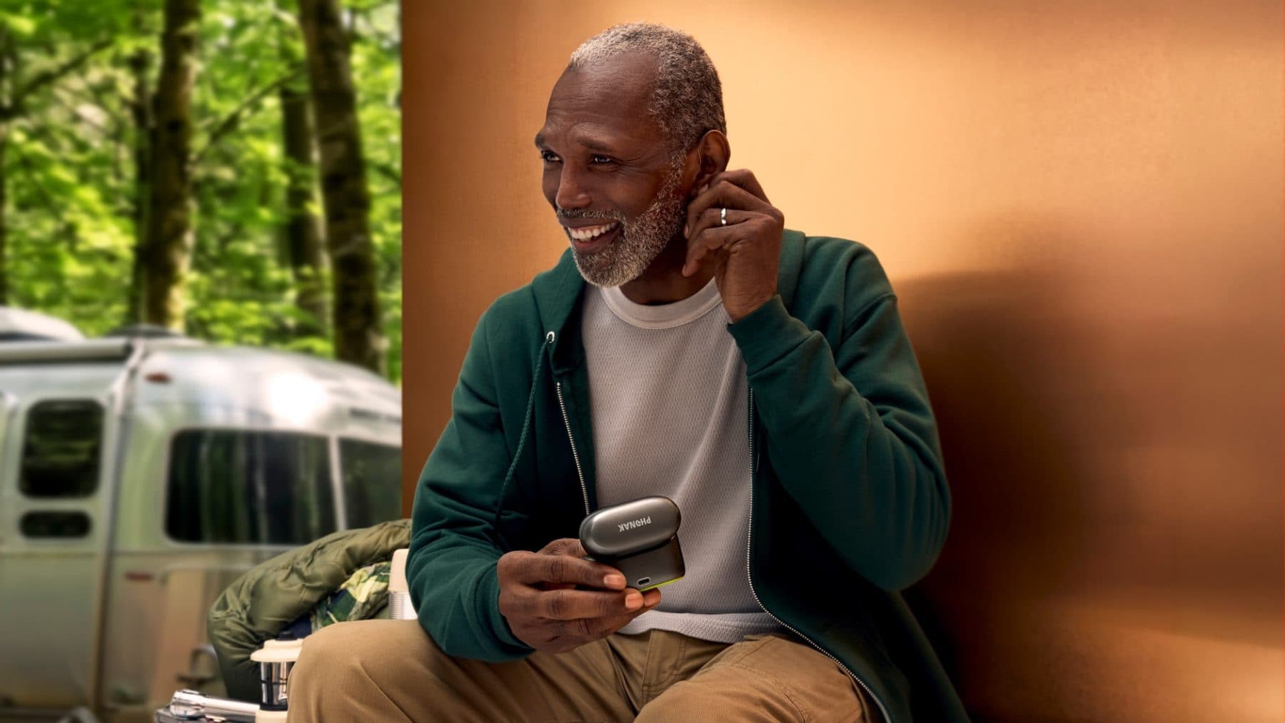 A smiling man at a campsite holding his Phonak hearing aid charging case